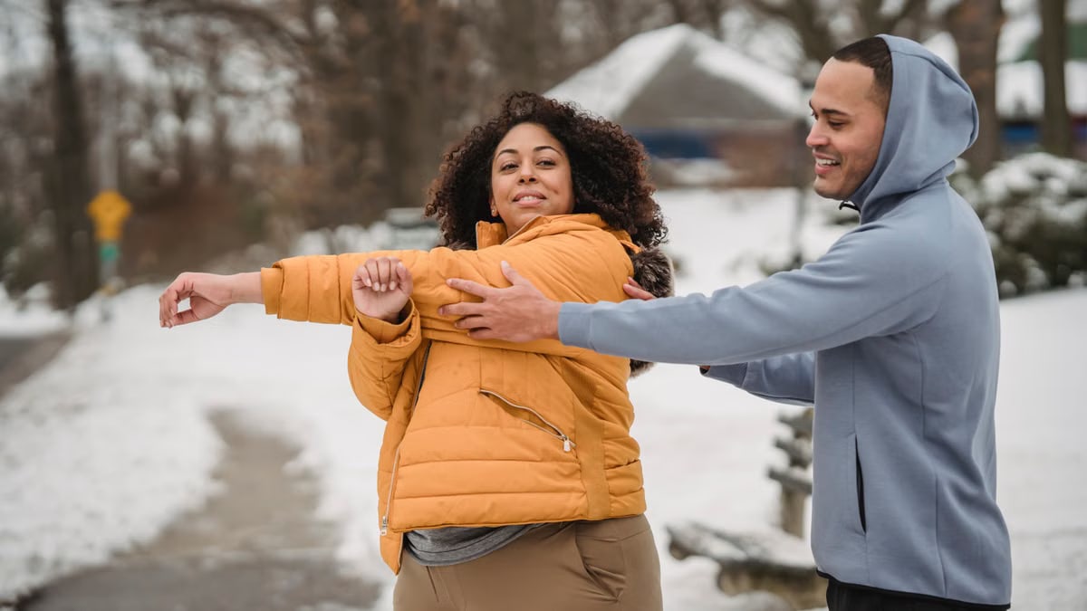 Positive African American overweight female in outerwear smiling while doing warming up exercise during personal training with coach